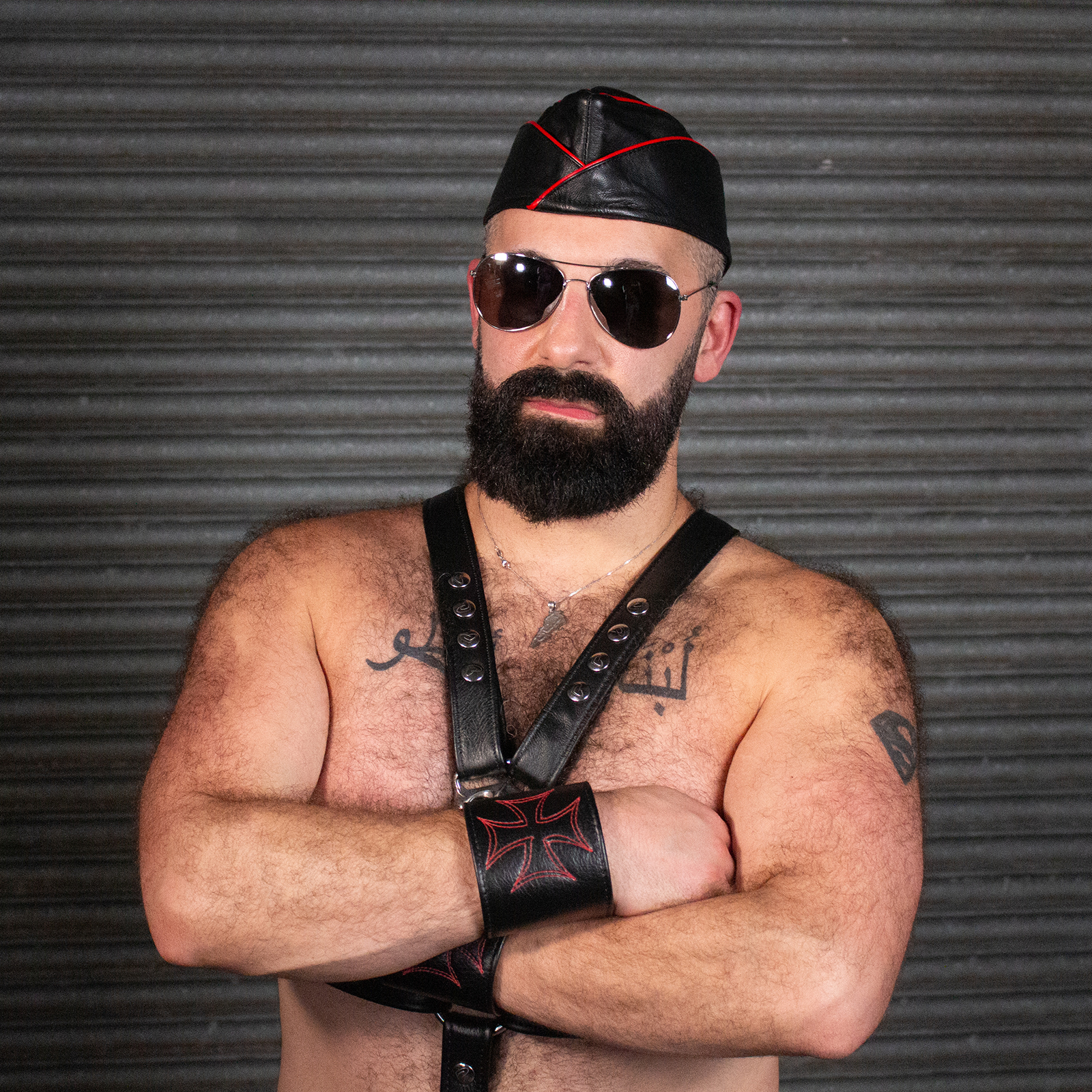 A sexy hairy man is wearing accessories including a garrison cap, a harness, leather wristbands, and aviator sunglasses.