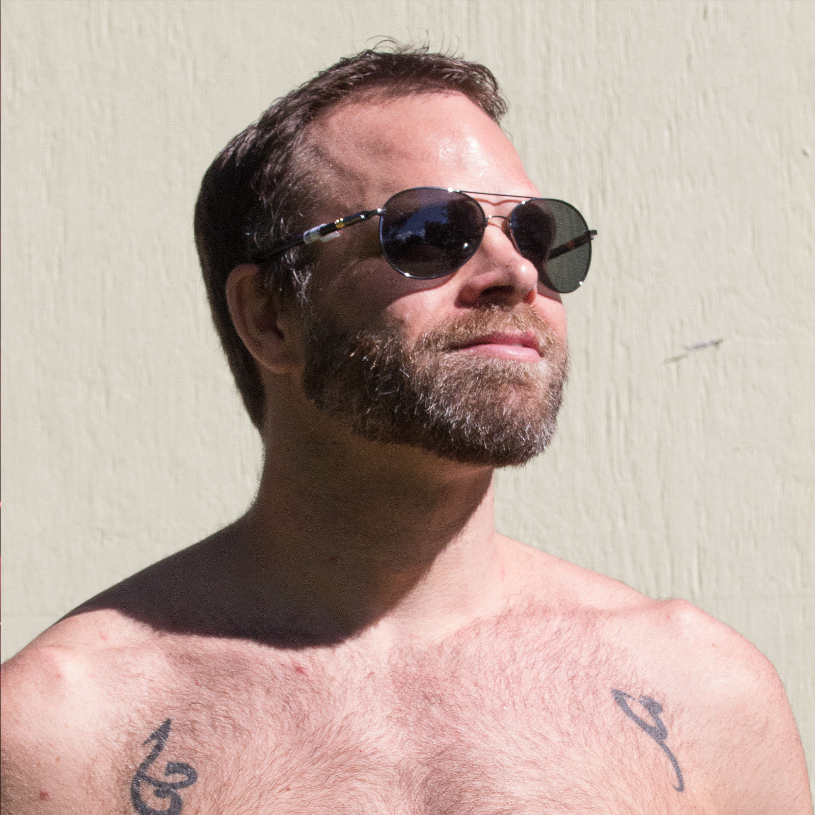 A bearded man stands in the sun, he bare chest getting a tan, good thing he’s wearing a pair of sexy aviator sunglasses.